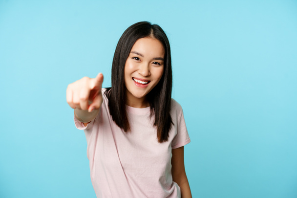 Sei tu. Sorridente felice donna asiatica puntando il dito contro la fotocamera, congratulandosi, invitando le persone, in piedi in t-shirt su sfondo blu - Foto, immagini