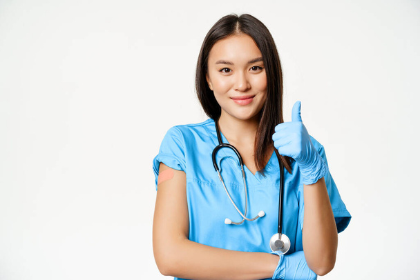 Hymyilevä sairaanhoitaja, aasialainen naislääkäri pensaikossa, näyttää peukalot ylös merkki ja rokotettu käsi lääkelaastilla, suosittelee rokotusta covid-19, valkoinen tausta - Valokuva, kuva