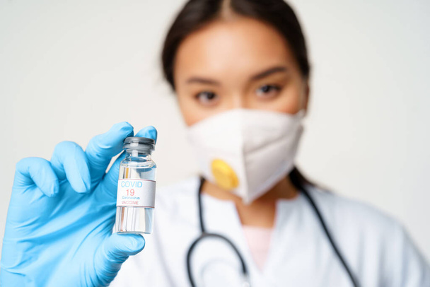 Concepto de vacunación y prevención covid-19. primer plano retrato de asiático médico en respirador muestra botella con coronavirus vacuna, de pie sobre fondo blanco - Foto, Imagen