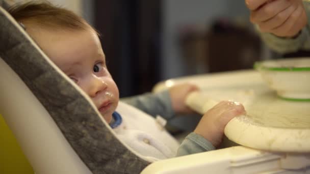 Kleine jongen zit in een kinderstoel en eet puree gemaakt van gepureerd fruit dat zijn moeder hem geeft - Video