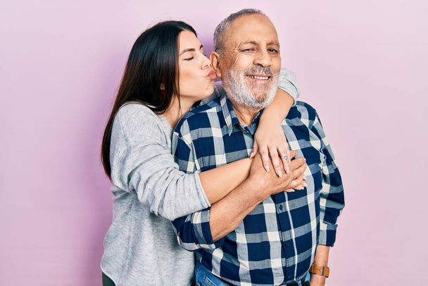 Young brunette woman and senior man standing over pink background. Daughter and father hugging and bonding together as happy family - Foto, Imagen