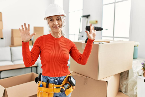 Femme aux cheveux gris d'âge moyen portant un casque debout à la nouvelle maison montrant et pointant vers le haut avec les doigts numéro dix tout en souriant confiant et heureux.  - Photo, image