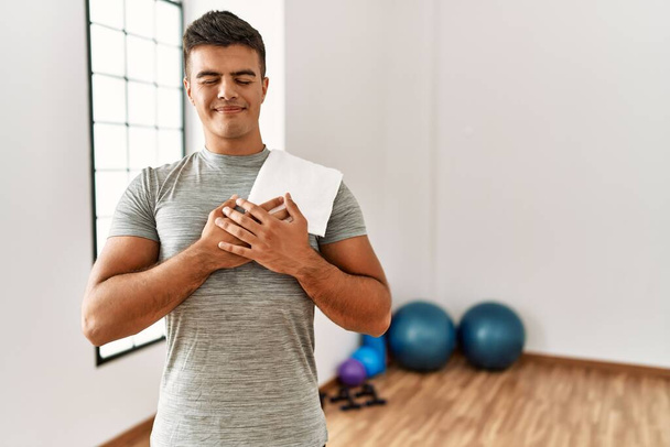 Young hispanic man wearing sportswear and towel at the gym smiling with hands on chest with closed eyes and grateful gesture on face. health concept.  - Foto, imagen