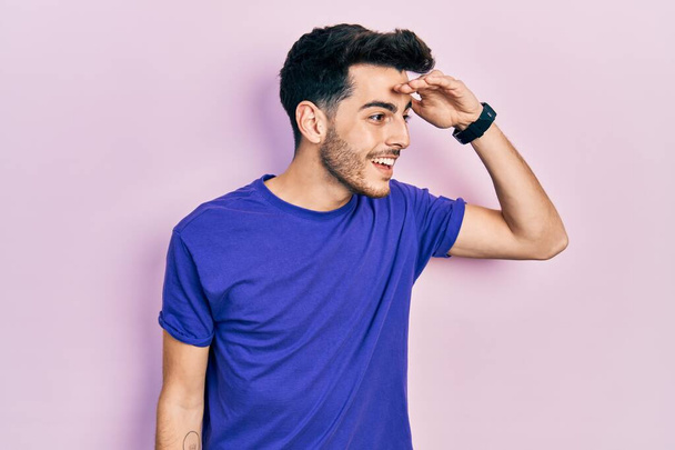 Joven hombre hispano vistiendo camiseta casual muy feliz y sonriente mirando lejos con la mano sobre la cabeza. concepto de búsqueda.  - Foto, Imagen
