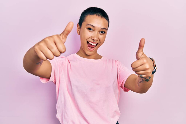 Красивая латиноамериканка с короткими волосами в обычной розовой футболке, одобряющая позитивный жест рукой, улыбается и радуется успеху. жест победителя.  - Фото, изображение