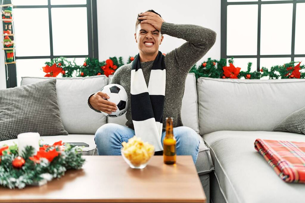 Young hispanic man football hooligan holding ball and beer stressed and frustrated with hand on head, surprised and angry face  - Φωτογραφία, εικόνα