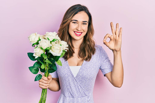 Νεαρό ισπανόφωνο κορίτσι κρατώντας λουλούδια κάνει ok υπογράψει με τα δάχτυλα, χαμογελώντας φιλικό gesturing εξαιρετικό σύμβολο  - Φωτογραφία, εικόνα