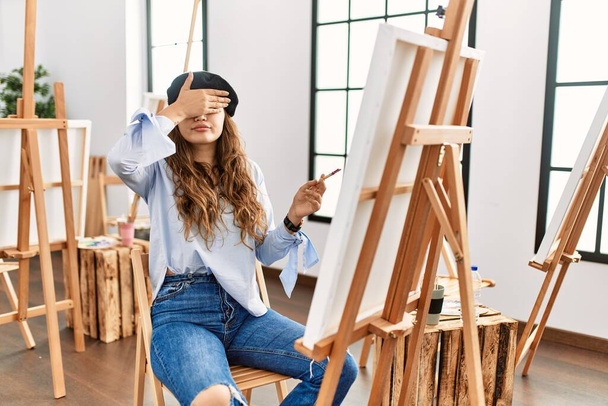 Młoda latynoska artystka malująca na płótnie w pracowni artystycznej, okrywająca oczy ręką, wyglądająca poważnie i smutno. Koncepcja niewidoczności, ukrywania i odrzucania  - Zdjęcie, obraz