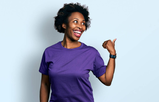 Африканская американка с афроволосами в повседневной фиолетовой футболке улыбается со счастливым лицом, глядя и указывая на сторону с большим пальцем вверх.  - Фото, изображение
