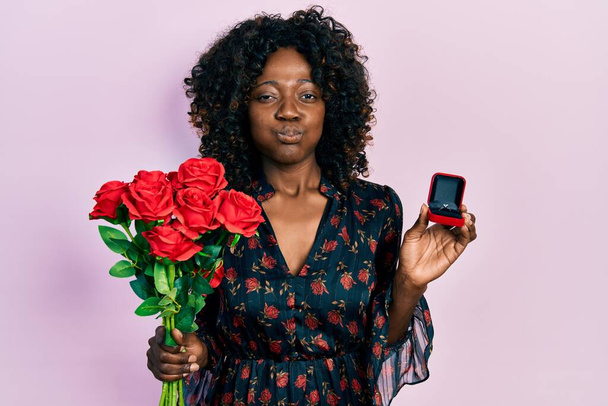 Young african american woman holding bouquet of flowers and engagement ring puffing cheeks with funny face. mouth inflated with air, catching air.  - Φωτογραφία, εικόνα