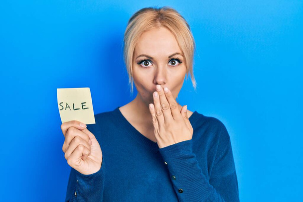 Young blonde woman holding sale word reminder covering mouth with hand, shocked and afraid for mistake. surprised expression  - Photo, Image