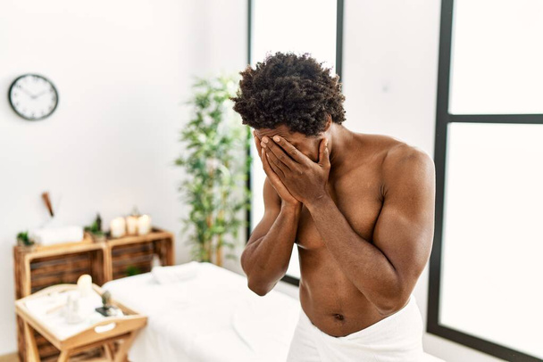 Fiatal afro-amerikai férfi félmeztelen törülközőben áll a szépségszalonban szomorú arckifejezéssel, miközben kezekkel takarja el az arcát, miközben sír. depresszió fogalma.  - Fotó, kép