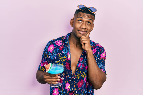 Young black man drinking tropical cocktail with alcohol serious face thinking about question with hand on chin, thoughtful about confusing idea  - 写真・画像