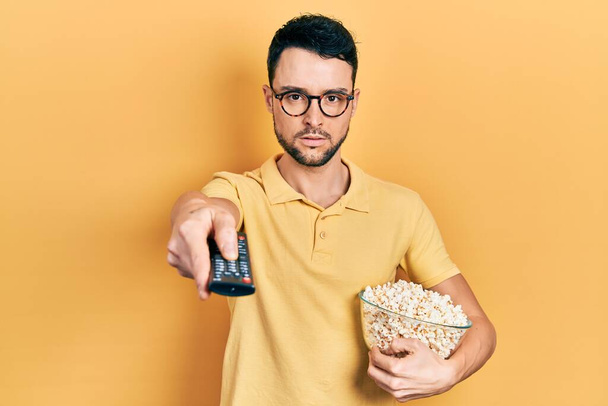 Giovane uomo ispanico mangiare popcorn con controllo tv scettico e nervoso, accigliato sconvolto a causa di un problema. persona negativa.  - Foto, immagini