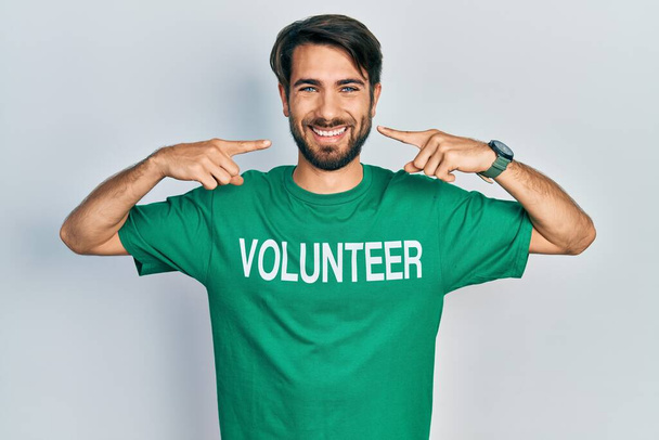 Junger hispanischer Mann im freiwilligen T-Shirt lächelt fröhlich und zeigt mit den Fingern Zähne und Mund. Zahngesundheitliches Konzept.  - Foto, Bild