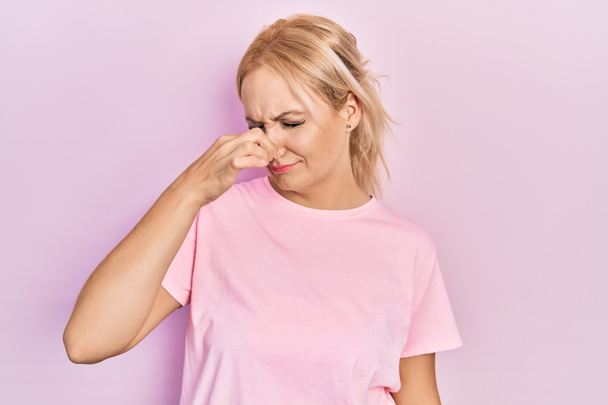 Jonge blonde vrouw draagt casual roze t-shirt ruiken iets stinkend en walgelijk, ondraaglijke geur, houden adem met vingers op de neus. slechte geur  - Foto, afbeelding