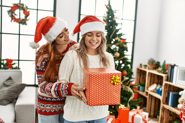 Woman couple hugging each other holding gift standing by christmas tree at home - Foto, Imagem