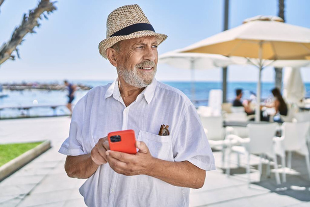 シニア男笑顔自信を持って着て夏の帽子を使用してスマートフォンで海辺で - 写真・画像