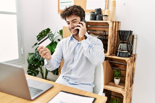 Jonge Spaanse zakenman in gesprek op de smartphone met een creditcard op kantoor. - Foto, afbeelding
