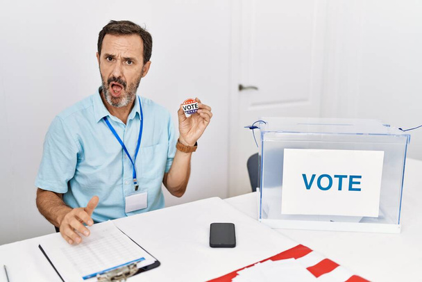 Hombre de mediana edad con barba sentado por votación sosteniendo voto insignia en la cara de choque, mirando escéptico y sarcástico, sorprendido con la boca abierta  - Foto, Imagen