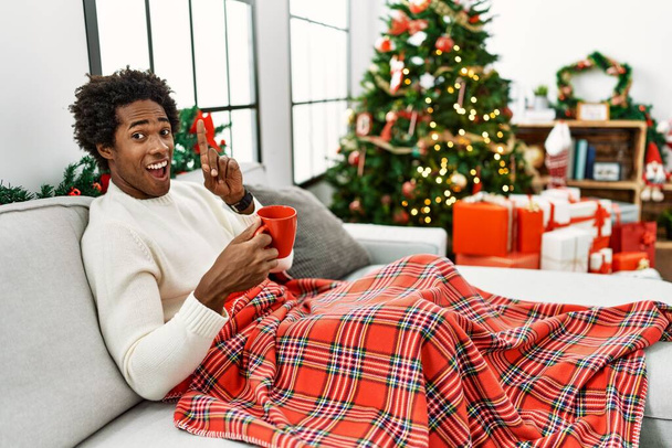 Fiatal afro-amerikai férfi ül a kanapén, kávét iszik karácsonyfán, ujjal mutogatva a sikeres ötletre. kilépett és boldog volt. Egyes számú..  - Fotó, kép