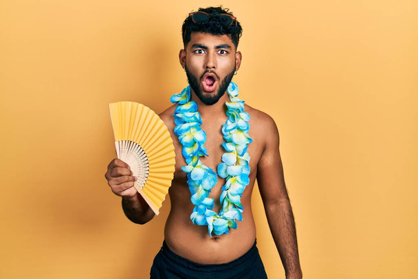 Arab man with beard wearing hawaiian lei holding handfan scared and amazed with open mouth for surprise, disbelief face  - Фото, изображение
