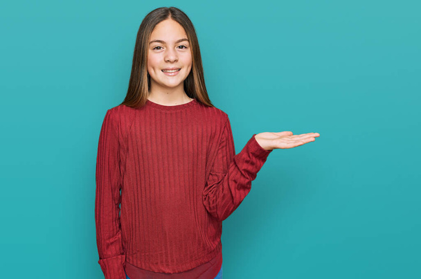 mooi brunette klein meisje dragen casual trui glimlachen vrolijk presenteren en wijzen met palm van de hand kijken naar de camera.  - Foto, afbeelding