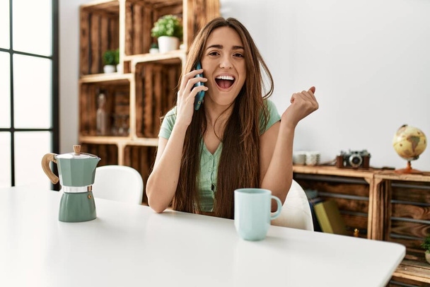 Νεαρή Ισπανίδα γυναίκα που μιλάει στο smartphone κάθεται στο τραπέζι στο σπίτι ουρλιάζοντας περήφανη, γιορτάζοντας τη νίκη και την επιτυχία πολύ ενθουσιασμένη με σηκωμένα χέρια  - Φωτογραφία, εικόνα