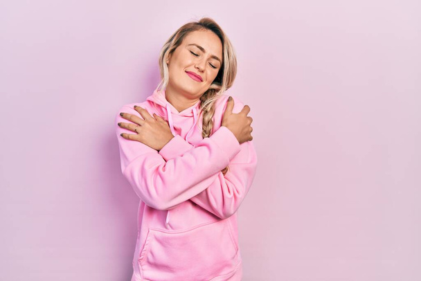 Hermosa mujer rubia joven con sudadera rosa abrazándose feliz y positivo, sonriendo confiado. amor propio y cuidado personal  - Foto, Imagen
