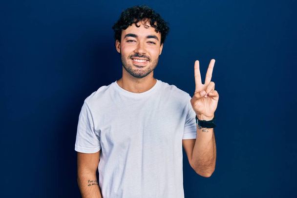 Young hispanic man wearing casual white t shirt smiling with happy face winking at the camera doing victory sign. number two.  - Photo, Image