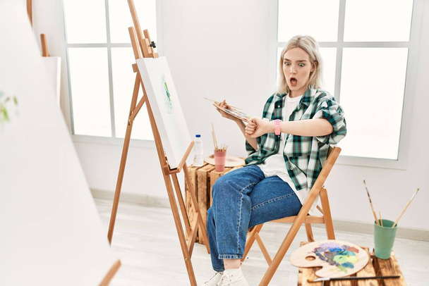 Young artist woman painting on canvas at art studio looking at the watch time worried, afraid of getting late  - Фото, изображение