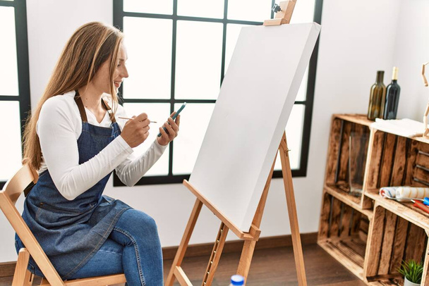 Nuori valkoihoinen nainen hymyilee luottavainen piirustus käyttäen älypuhelin taidestudiossa - Valokuva, kuva