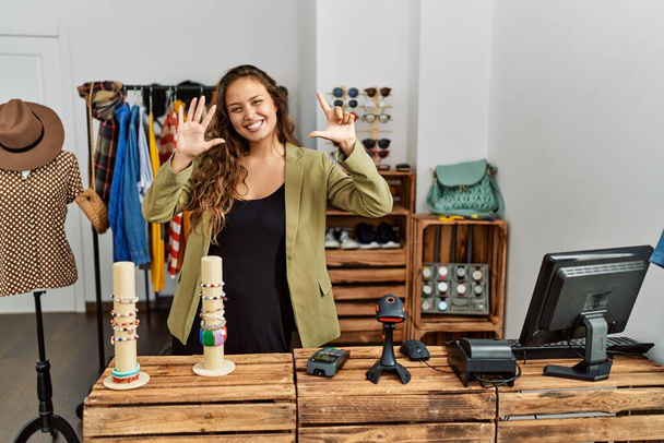 Beautiful hispanic woman working at fashion shop showing and pointing up with fingers number seven while smiling confident and happy.  - Photo, Image