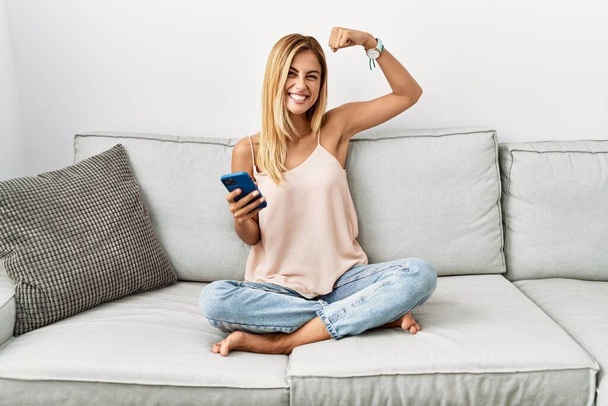 Blonde beautiful young woman sitting on the sofa at home using smartphone strong person showing arm muscle, confident and proud of power  - Foto, Imagen
