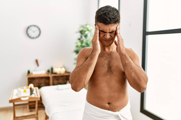 Young hispanic man standing shirtless at spa center with hand on head for pain in head because stress. suffering migraine.  - Fotografie, Obrázek