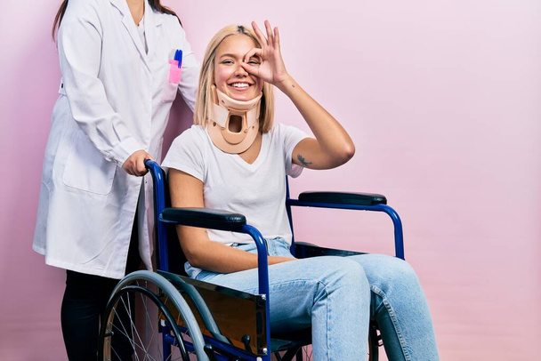 Beautiful blonde woman sitting on wheelchair with collar neck doing ok gesture with hand smiling, eye looking through fingers with happy face.  - Fotografie, Obrázek