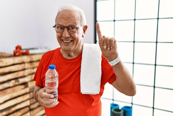 Älterer Mann in Sportbekleidung und Handtuch in der Turnhalle lächelt erstaunt und überrascht und zeigt mit erhobenen Fingern nach oben.  - Foto, Bild