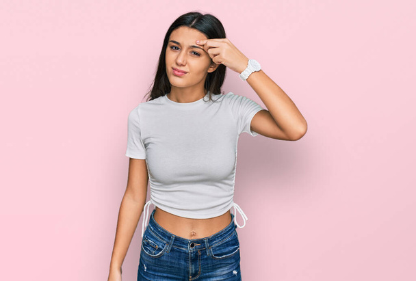 若いヒスパニック系の女の子の額ににきび、黒頭の醜い感染に不幸なポインティングカジュアルな白いTシャツを着て。にきびや皮膚の問題  - 写真・画像