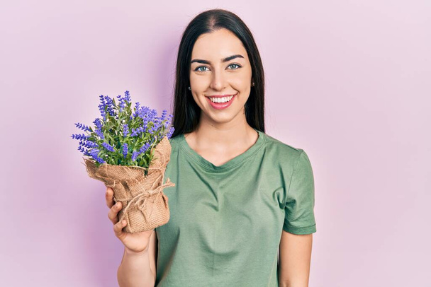 Gyönyörű nő kék szemekkel gazdaság levendula növény néz pozitív és boldog álló és mosolygó magabiztos mosollyal mutatja fogak  - Fotó, kép