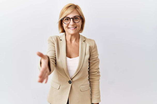 Middle age blonde business woman standing over isolated background smiling friendly offering handshake as greeting and welcoming. successful business.  - Fotó, kép