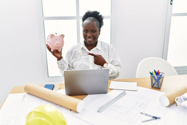 Jovem africana trabalhando no estúdio de arquitetura segurando porquinho banco sorrindo feliz apontando com a mão e o dedo  - Foto, Imagem