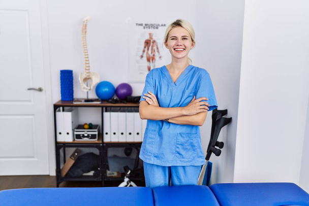 Jonge fysiotherapeut vrouw die werkt bij pijn herstel kliniek blij gezicht glimlachen met gekruiste armen kijken naar de camera. positieve persoon.  - Foto, afbeelding