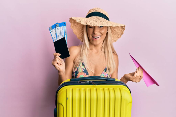 Молода блондинка в бікіні тримає паспорт і паперовий літак підморгнувши дивиться на камеру з сексуальним виразом, веселим і щасливим обличчям
.  - Фото, зображення