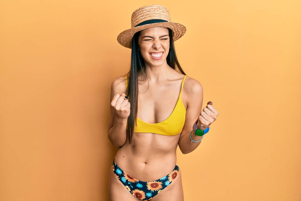 Junge hispanische Frau mit Bikini und Sommermütze, aufgeregt für den Erfolg, mit erhobenen Armen und geschlossenen Augen, die den Sieg lächelnd feiert. Siegerkonzept.  - Foto, Bild