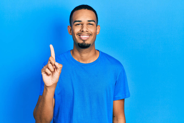 Jeune homme afro-américain portant un t-shirt bleu décontracté montrant et pointant vers le haut avec le doigt numéro un tout en souriant confiant et heureux.  - Photo, image