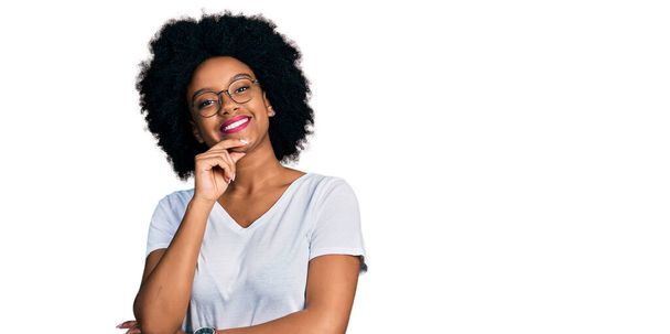 Joven mujer afroamericana con camiseta blanca casual mirando confiada a la cámara sonriendo con los brazos cruzados y la mano levantada en la barbilla. pensar positivamente.  - Foto, Imagen