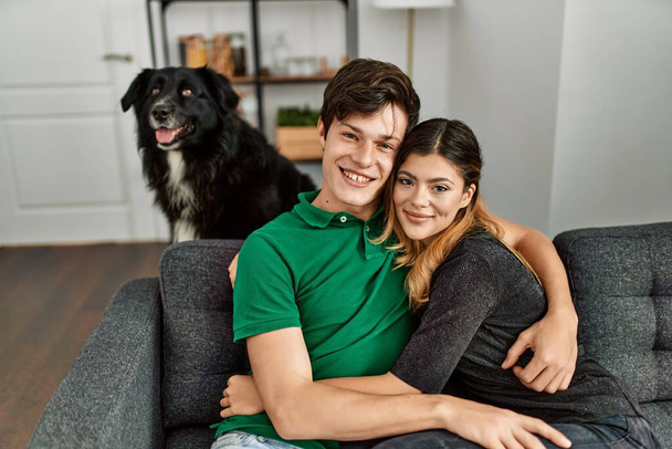 Junges kaukasisches Paar lächelt glücklich und umarmt sich auf dem Sofa sitzend mit Hund zu Hause. - Foto, Bild