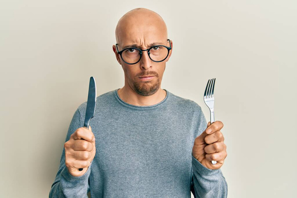 Homme chauve avec barbe tenant fourchette et couteau prêt à manger sceptique et nerveux, fronçant les sourcils bouleversés à cause de problèmes. personne négative.  - Photo, image