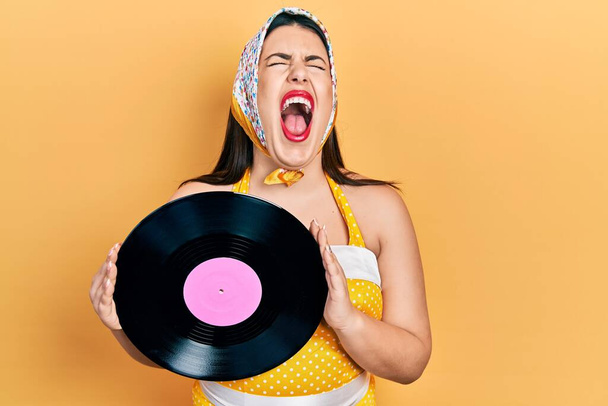 Young hispanic woman wearing pin up style holding vinyl disc angry and mad screaming frustrated and furious, shouting with anger looking up.  - Photo, Image