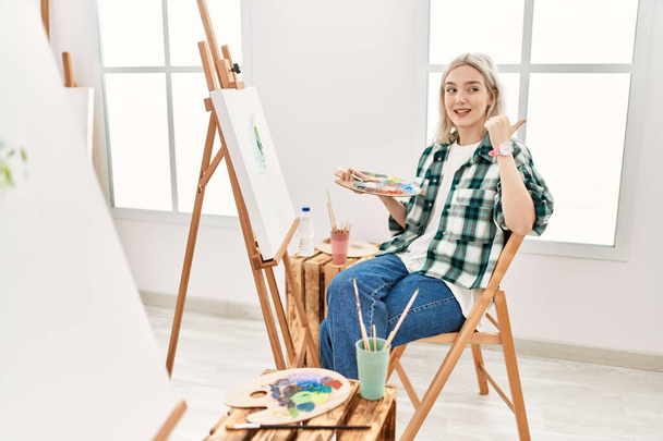 Молода художниця малює на полотні в художній студії, усміхаючись із щасливим обличчям, дивлячись і вказуючи на бік з великим пальцем вгору
.  - Фото, зображення